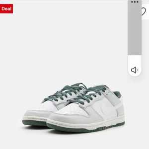 Nike Dunk Low vit/grön skinn - Nike Dunk Low sneakers i vitt och ljusgrått skinn med gröna detaljer och gröna snören. Klassisk siluett med perforerad tå och platt sula. Perfekta för dig som gillar streetstyle och vill ha en clean look.