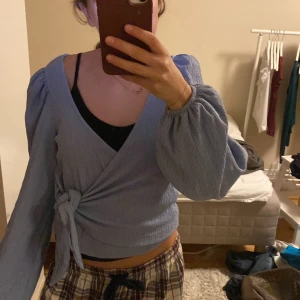 Ljusblå blus från Monki - Supersöt ljusblå omlottopp från Monki med långa puffärmar och knyt i sidan. Toppen har en ribbad struktur och croppad passform som ger en trendig vibe. Perfekt att styla med jeans eller kjol för en chill look.