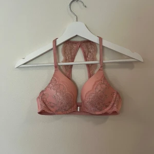 Rosa spetsig bh från Hunkemöller - Supersöt rosa bh från Hunkemöller med glittriga spetsdetaljer och snygg racerback i spets. Toppen har vadderade kupor och smidig knäppning framtill. Perfekt för dig som gillar feminina och trendiga detaljer.