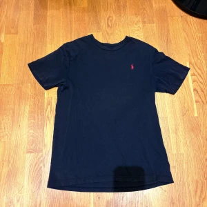 Svart t-shirt från Ralph Lauren - Klassisk svart t-shirt från Ralph Lauren med röd broderad logga på bröstet. T-shirten har rund halsringning och korta ärmar. Perfekt basic-plagg med snygg passform och diskret branding.