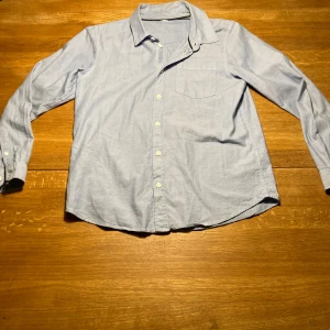 Ljusblå bomullsskjorta med ficka - En klassisk ljusblå skjorta i bomull med lång ärm, bröstficka och knappar framtill. Skjortan har en enkel och stilren design med normal passform och mjuk känsla. Perfekt för dig som gillar en clean och avslappnad look.