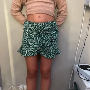 Grön blommig volangkjol med rosett - Supersöt grön kortkjol med vitt blommönster och volangkant. Kjolen har en omlott-look med en fin rosett i sidan och volanger längs nederkanten. Perfekt för vår och sommar när du vill ha en lekfull och trendig stil.