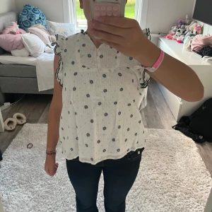 Vit blommig blus med volangärm - Supersöt vit blus från Lindex med blommor i mörkblått. Blusen har volangärmar med mörkblå kanter, små knappar framtill och lätt v-ringning. I storlek 158/164.