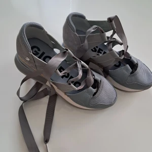 Grå sandaler från Gioseppo med snörning - Snygga grå sandaler från Gioseppo med breda band och långa grå snören att knyta runt vristen. Ovandel i mocka och textil, detaljer i skinnimitation och mesh. Sula med flätad jutedetalj och vit plattform. Perfekt för dig som gillar en sportig men trendig look.