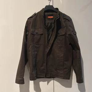 Mörkgrön militärinspirerad jacka - Snygg mörkgrön jacka med coola broderade patchar på ärm och bröst. Jackan har axelklaffar, två bröstfickor med knappar och dragkedja framtill. Tillverkad i bomull och har en klassisk militärlook som sticker ut.