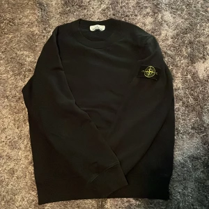 Svart sweatshirt från Stone Island - Svart sweatshirt från Stone Island med klassisk rund hals och ikonisk logotyp-patch på vänster ärm. Tröjan har en clean look och är tillverkad i mjuk bomull, perfekt för en stilren och avslappnad vibe.