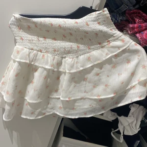 Vit blommig volangkjol - Supersöt vit kortkjol med små rosa blommor och volanger. Kjolen har smock i midjan för en bekväm passform och är gjord i ett lätt, luftigt tyg. Perfekt för varma dagar när du vill känna dig extra gullig.