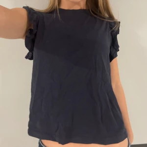 Mörkblå volangtopp med kort ärm - Snygg mörkblå topp med volangdetaljer på ärmarna och rak passform. Toppen är kortärmad och tillverkad i mjuk bomull, perfekt för en chill och stilren look. Enkel att matcha med jeans eller kjol för en fin look🤍