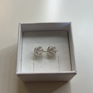 Snygga silvriga örhängen med knutdesign - Ett par stilrena silvriga örhängen i form av små knutar. De har en glittrig detalj i mitten som ger extra bling. Perfekta för dig som gillar minimalistiska smycken med en twist. Kommer i en söt ask.
