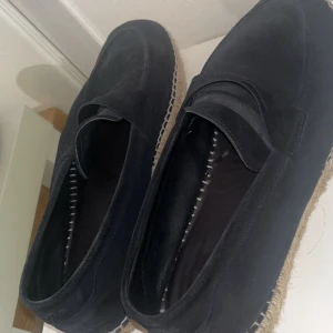 Svarta mockaloafers med repdetalj - Snygga svarta loafers i mocka med klassisk pennyloafers-design och grov sula i repstil. Perfekta för dig som vill ha en stilren men chill sko till våren eller sommaren. Tidlös look med rund tå och platt sula.