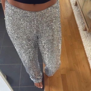 Glittriga silverbyxor med paljetter - Säljer ett par sjukt snygga byxor i silver med massor av paljetter som glittrar i ljuset. Byxorna har hög midja och ett rakt, lite loose fit. Perfekta om du vill sticka ut och glänsa på nästa event. Materialet känns mjukt och följsamt mot huden.