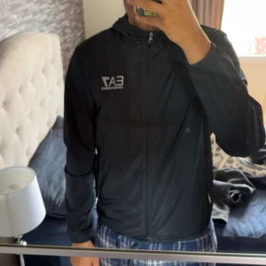 Svart vindjacka från EA7 Emporio Armani - Svart vindjacka från EA7 Emporio Armani med huva och dragkedja framtill. Kommer inte till användning längre 