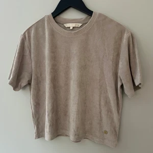 Beige frotté t-shirt Pernilla Wahlgren - Supermjuk beige t-shirt från Pernilla Wahlgren i frotté. Klassisk rund hals, korta ärmar och relaxed fit. Snygg detalj med liten guldfärgad ananas vid nederkanten. Perfekt för chill dagar eller när du vill ha något bekvämt men ändå stilrent.