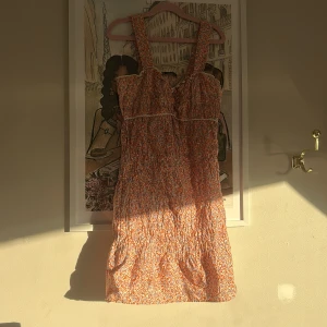 Blommig kort klänning från Zara - Supersöt kort klänning från Zara med blommigt mönster i orange, lila och beige. Klänningen har breda axelband, smockad överdel och volangdetaljer vid bysten. Perfekt för varma dagar och ger en riktigt somrig vibe.