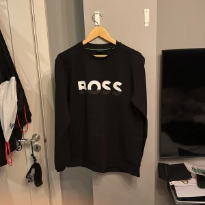 Svart sweatshirt från Boss - Snygg svart sweatshirt från Boss med deras logga i vitt och grått på bröstet. Tröjan har långa ärmar och en klassisk rund halsringning. Perfekt för en stilren och avslappnad look.