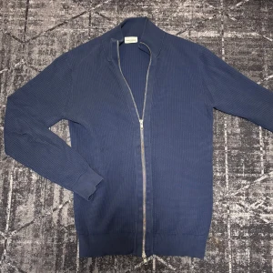 Blå Full Zip Kofta - Snygg blå kofta från John Henric med dragkedja framtill. Den har en ribbad krage och är tillverkad i ett bekvämt material. Perfekt för en stilren look.