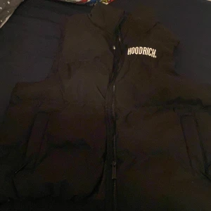 Svart dunväst från Hoodrich - Säljer en svart dunväst från Hoodrich med dragkedja framtill. Västen har en stilren design med Hoodrich-loggan tryckt på bröstet och ryggen. Perfekt för att hålla värmen med stil. M men passar även S