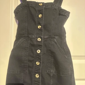 Svart jeansklänning med knappar - Trendig svart jeansklänning med knappar framtill och justerbara axelband. Perfekt för en avslappnad stil med praktiska fickor. Enkla detaljer som ger en cool look.