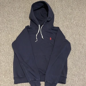 Mörkblå hoodie från Ralph Lauren - Säljer en stilren mörkblå hoodie från Ralph Lauren med en liten röd logga på bröstet. Tröjan har en klassisk känguruficka och justerbar huva med vita snören. Perfekt för en avslappnad stil.