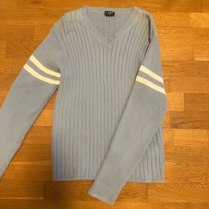 Ljusblå ribbad tröja från Logan - Säljer en stilren ljusblå ribbad tröja från Logan med vita ränder på ärmarna. Tröjan har en v-ringad hals och långa ärmar, perfekt för en avslappnad look. Passar bra till jeans eller kjol. Hör av dig vid intresse!