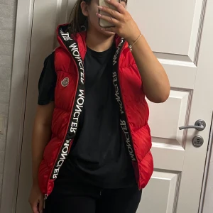 Röd dunväst från Moncler - Snygg röd dunväst från Moncler med dragkedja och huva. Västen har märkets logga på bröstet och text längs dragkedjan. Perfekt för kyligare dagar när du vill hålla stilen. Denna väst har aldrig använda den är helt ny och är i strl 15-16 men osssar även xs och s! Säljer denna för 650 kr!