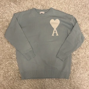 Ljusblå stickad tröja från Ami - Säljer en stilren ljusblå stickad tröja från Ami med ett vitt hjärtmönster och bokstaven A på bröstet. Tröjan har långa ärmar och ribbade muddar vid ärmslut och nederkant. Perfekt för en avslappnad och trendig look.