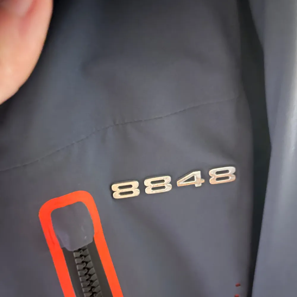 Säljer en svart jacka från 8848 Altitude med orange foder och coola märken på ärmen. Jackan har en dragkedja framtill och är perfekt för utomhusaktiviteter. Den har en sportig look med långärmade ärmar och en skön passform. Termobyxor medföljer, de är hel orange och har hängslen.. Takit.