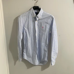 Rutig skjorta från Ralph Lauren - Snygg rutig skjorta från Ralph Lauren i blått och vitt. Skjortan har en slim fit passform och är långärmad med knappar framtill. Den har en broderad logotyp i rosa på bröstet. Perfekt för en stilren look!