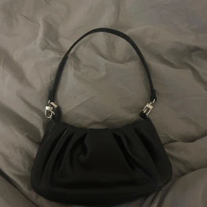 Svart axelväska med silverdetaljer - Snygg svart axelväska med stilrena silverdetaljer. Perfekt för att bära dina nödvändigheter med stil. Väskan har en enkel design som passar till många olika outfits.