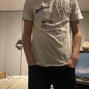 Vit t-shirt med blått tryck - Säljer en vit t-shirt med ett stilrent blått tryck som föreställer solstolar och parasoll. Perfekt för en avslappnad stil. 