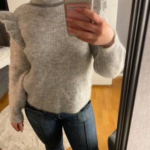 Grå stickad tröja  - En grå stickad tröja med volanger på axlarna och en polokrage. Är använd få antal gånger och i väldigt bra skick.