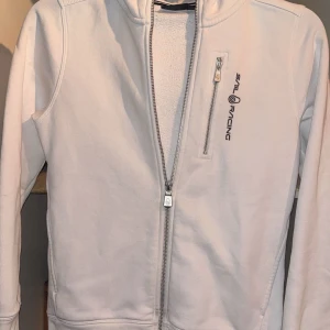Vit tröja  från Sail Racing - Snygg vit tröja  från Sail Racing med dragkedja och en bröstficka med logotyp. Perfekt för en stilren look. Tröjan har långa ärmar och en enkel design som passar till mycket.