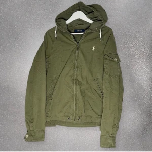 Ralph lauren jacka - Snygg grön jacka med huva från Polo Ralph Lauren. Jackan har dragkedja framtill och en broderad logga på bröstet. Den har långa ärmar och justerbara muddar. Perfekt för en avslappnad stil.