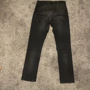 Svarta jeans från Nudie Jeans - Snygga mörk gråa jeans från Nudie Jeans med en klassisk femficksdesign med en diskret logga där bak. De är i väldigt bra skick då de bara har använts fyra gånger. Jeansen sitter slim och är perfekta för en stilren look. Priset är ej hugget i sten och går att diskutera.