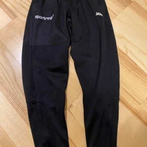 Svarta träningsbyxor från Jako - Svarta träningsbyxor från Jako med elastisk midja och logga på låret. Perfekta för sport och aktivitet, tillverkade i ett bekvämt syntetmaterial som ger bra rörelsefrihet.