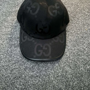 Svart keps med GG-mönster - Snygg svart keps från Gucci med det ikoniska GG-mönstret. Kepsen har en justerbar rem baktill för perfekt passform och är tillverkad i ett slitstarkt material. Perfekt för att ge din outfit en lyxig touch.