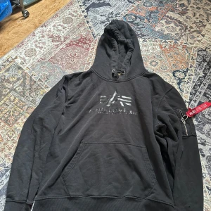 Svart hoodie från Alpha Industries - Säljer en svart hoodie från Alpha Industries med deras ikoniska logga på bröstet. Hoodien har en praktisk ficka framtill och en dragkedja med röd detalj på ärmen. Perfekt för en casual look.        (Går att pruta!!)