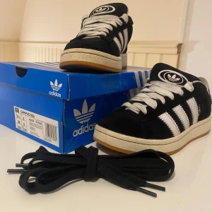 Svarta Adidas Campus 00s - Snygga svarta äkta Adidas Campus sneakers med vita detaljer och klassiska tre ränder på sidan. Skorna kommer med vita skosnören och extra svarta skosnören. Perfekta för en stilren look. Skicket är helt okej. Nypris 1500. Är även öppen för byten.
