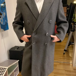 Kvalitets kappa i ull - Klassisk kvalitets kappa med dubbelknäppning. Den har en elegant design med breda slag och en rak passform. Karln på bilden har vanligtvis strl M.