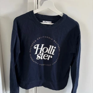 Mörkblå tröja från Hollister - Säljer en mörkblå tröja från Hollister med tryck i vitt på framsidan. Tröjan har långa ärmar och en rund halsringning. Perfekt för en avslappnad stil. Storlek S