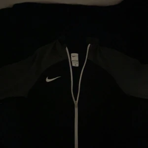   Nike zip - Säljer en svart och grå träningsjacka från Nike med Dri-FIT-teknologi. Jackan har en dragkedja framtill och är långärmad. Perfekt för träning och sportiga aktiviteter. Slim fit för en snygg passform. Den är helt ny