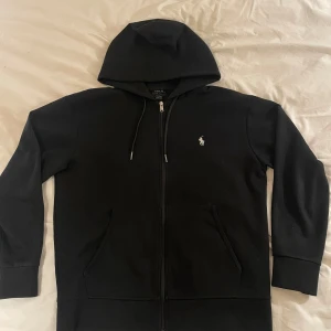 Svart hoodie från Ralph Lauren - Säljer en klassisk svart hoodie från Ralph Lauren med dragkedja och huva. Den har en liten broderad logga på bröstet och är tillverkad i ett mjukt material. Perfekt för en avslappnad stil. Pris kan diskuteras 