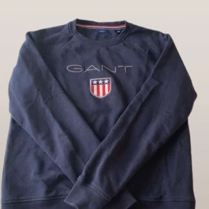 Mörkblå tröja från GANT - Säljer en mörkblå tröja från GANT med broderad logotyp och sköld på bröstet. Tröjan har långa ärmar och rund halsringning. Perfekt för en avslappnad stil. Pris kan diskuteras…😄