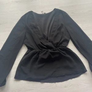 Svart omlottblus  - Snygg svart omlott topp, är lite mer ”genomskinlig” vid ärmarna och är urringad med stretchband i midjan. 