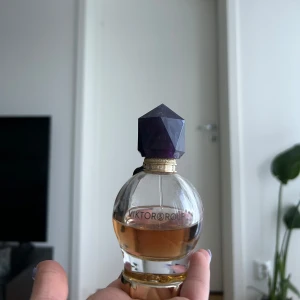 Parfym från Viktor & Rolf - Elegant parfymflaska från Viktor & Rolf med en unik, geometrisk lila kork. Flaskan är rund och genomskinlig med en gyllene vätska inuti. Perfekt för den som söker en lyxig doftupplevelse. 50% kvar! Nypris 1000kr