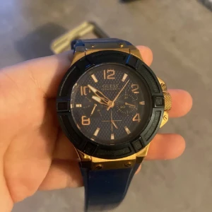 Blå och guld klocka från Guess - Snygg klocka från Guess med en blå urtavla och guld detaljer. Klockan har en robust design med en blå rem och tydliga siffror. Den är vattentålig upp till 100 meter och har en rostfri stålbaksida. Perfekt för den som vill ha en stilren och funktionell accessoar. Låda ingår in i köpen!
