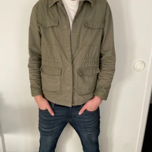 overshirt - Säljer en stilren olivgrön overshirt med långa ärmar och knappar framtill. Jackan har två bröstfickor och två större fickor nedtill, perfekt för en avslappnad look. Overshirten är i mycket bra skick nästan inte använd 