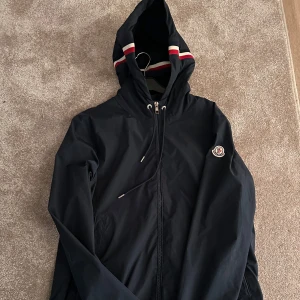 Svart vindjacka med huva - Snygg svart vindjacka från Moncler med huva och dragkedja. Jackan har en broderad logotyp på bröstet och snörning vid huvan. Perfekt för blåsiga dagar.