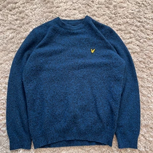 Blå stickad tröja från Lyle & Scott - Säljer en snygg blå stickad tröja från Lyle & Scott med deras ikoniska gula logotyp på bröstet. Tröjan har långa ärmar och en rund halsringning, perfekt för kyligare dagar. Passar både till jeans och chinos! Defekt är att den är nopprig