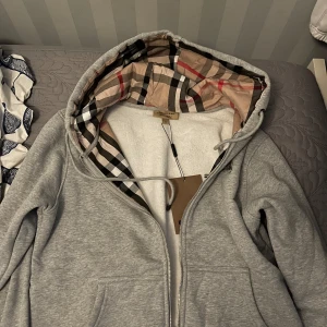 Grå huvtröja från Burberry - Säljer en stilren grå huvtröja från Burberry med klassiskt rutigt mönster i huvan. Tröjan har dragkedja framtill och praktiska fickor. Perfekt för en avslappnad look.
